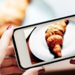 Mujer tomando fotografía de un croissant para su contenido en redes sociales.